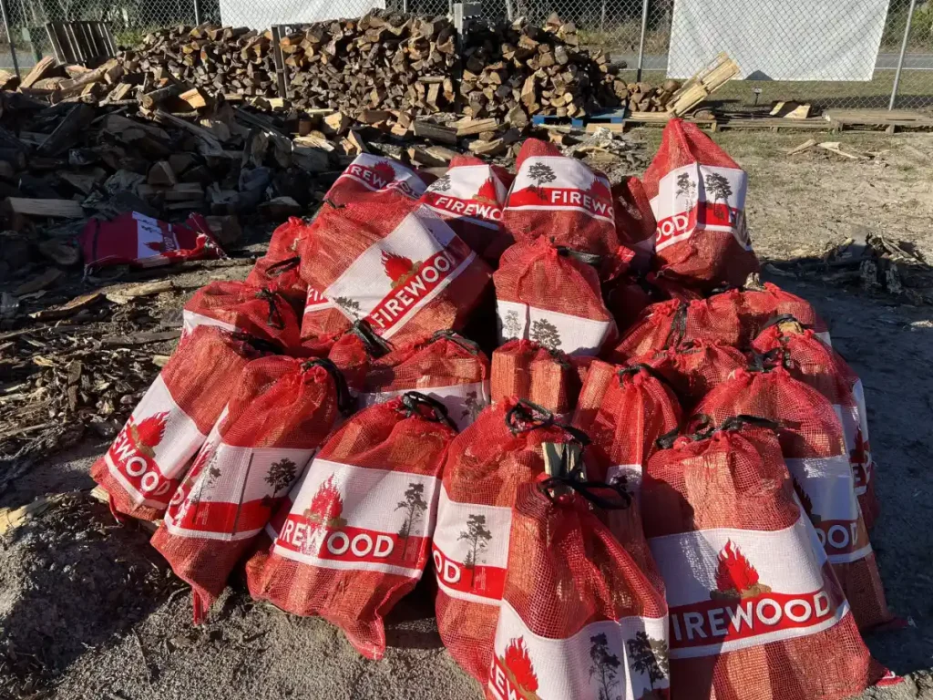 A pile of red mesh bags labeled "FIREWOOD" filled with chopped firewood is stacked outdoors in Sumter, FL, with loose firewood and a fence visible in the background.