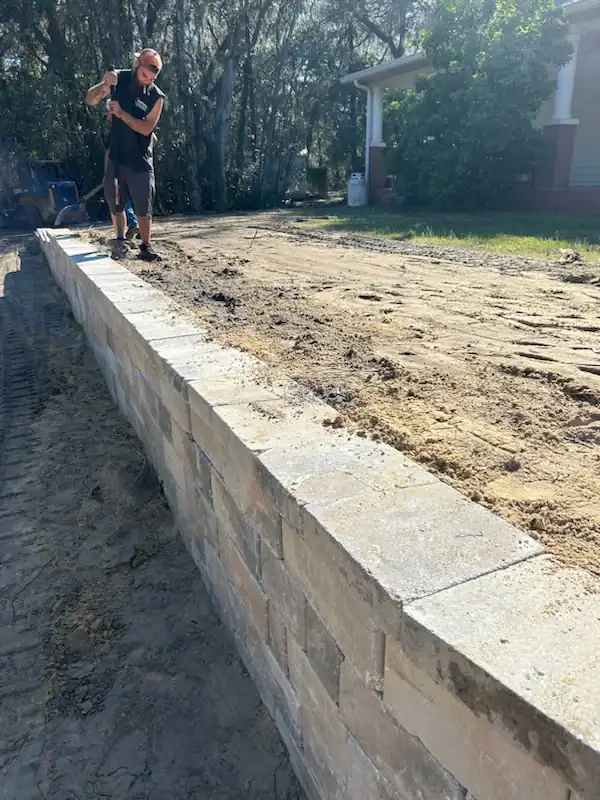 A landscaper from Hernando County uses a rake to level dirt on top of a newly built stone retaining wall next to a house, with trees visible in the background.