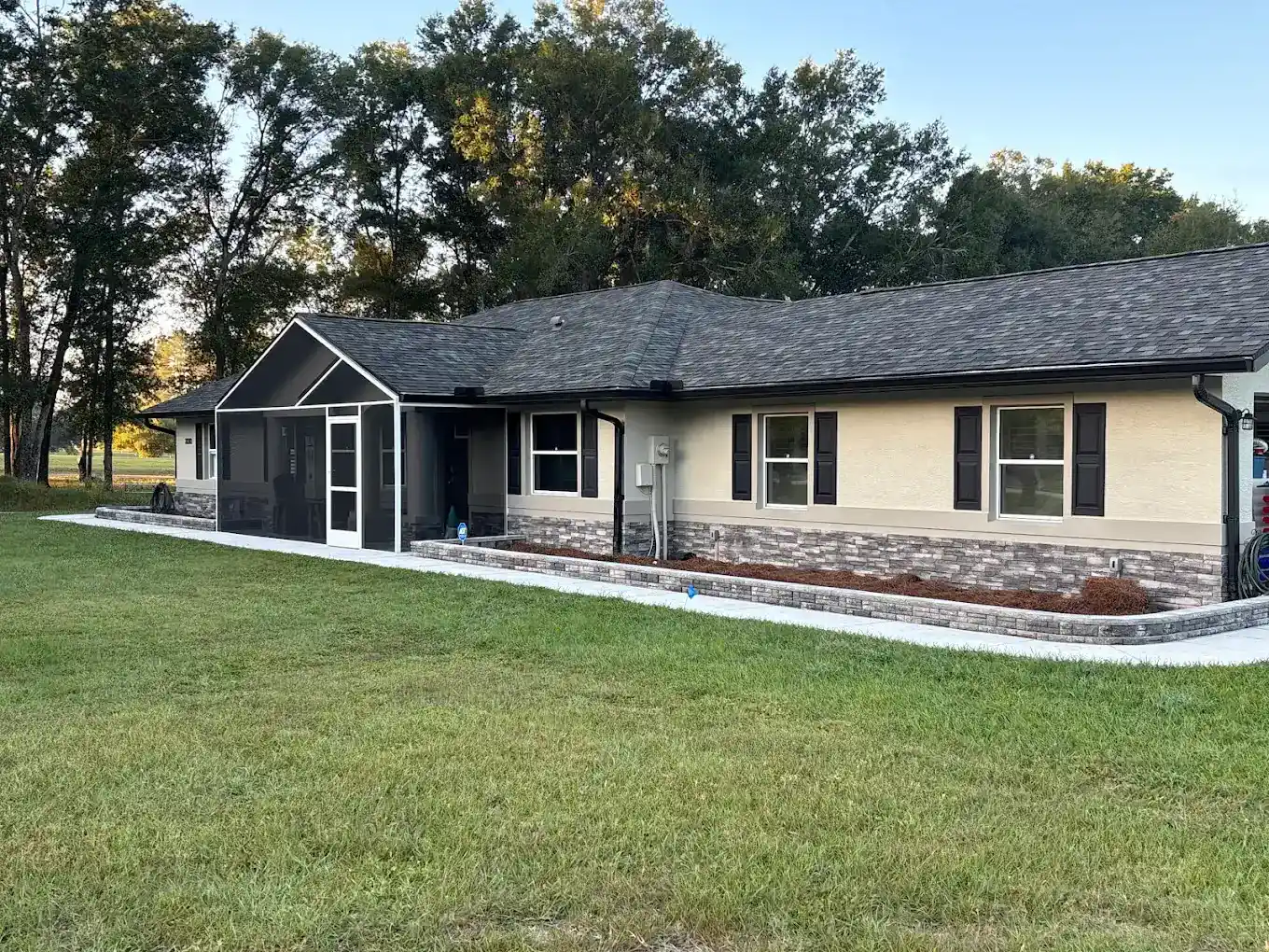 Single-story house in Hernando County, FL, with beige walls, dark brown shutters, and a screened-in porch. A wide, paved sidewalk and a grassy lawn sit in front, framed by trees in the background.