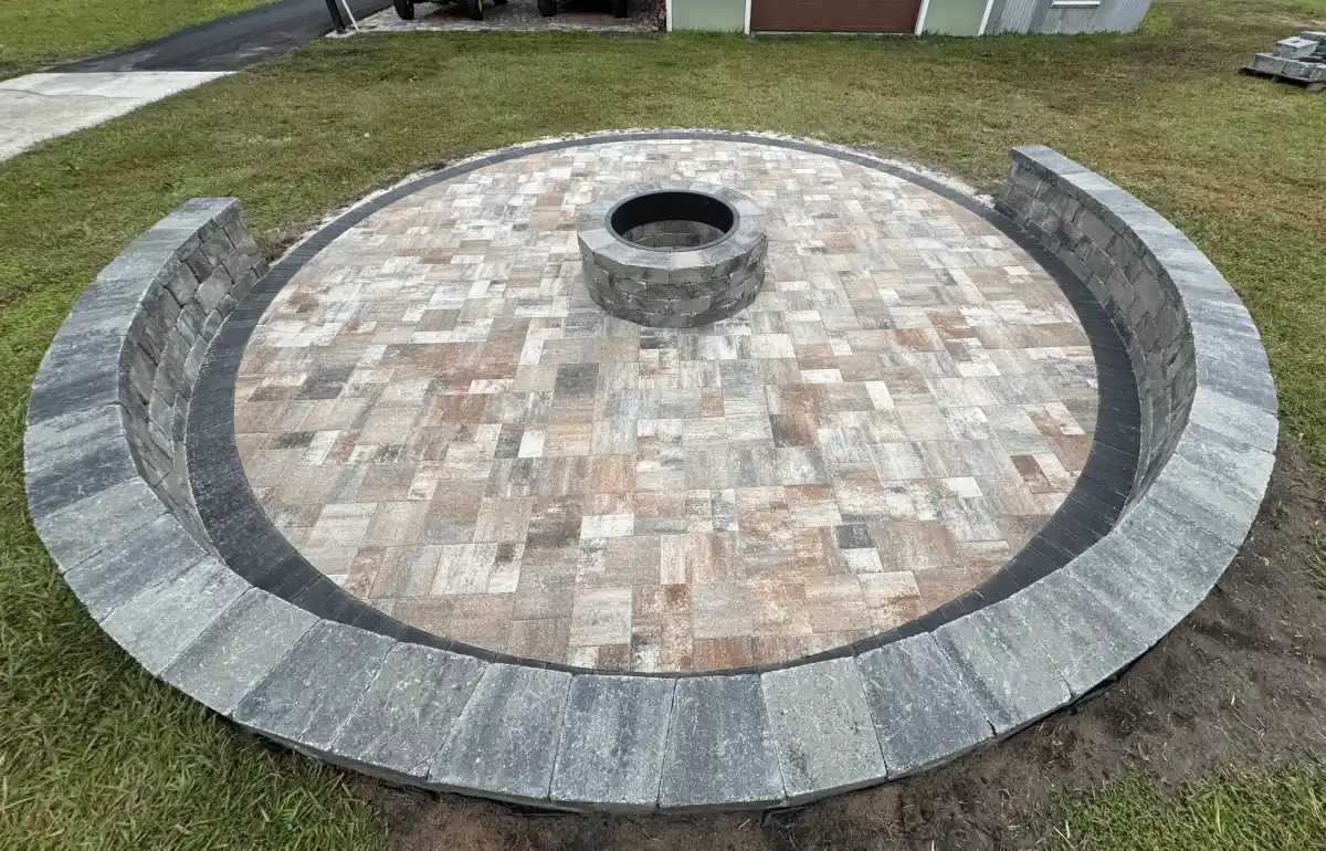 A circular stone patio with a built-in fire pit in the center, designed by Landscaper Citrus, features curved seating walls and sits on a grassy Sumter, FL lawn. The earth-toned pavers are set in square and rectangular patterns.