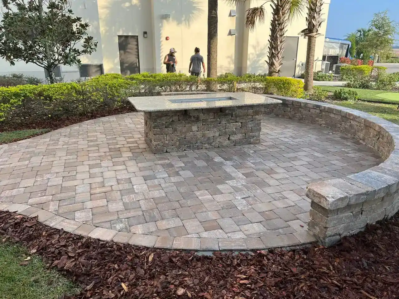 A circular stone patio with a built-in stone table and curved bench, designed by a skilled Hernando County landscaper, is surrounded by landscaped mulch and bushes; two people walk in the background near a light-colored FL building and palm trees.