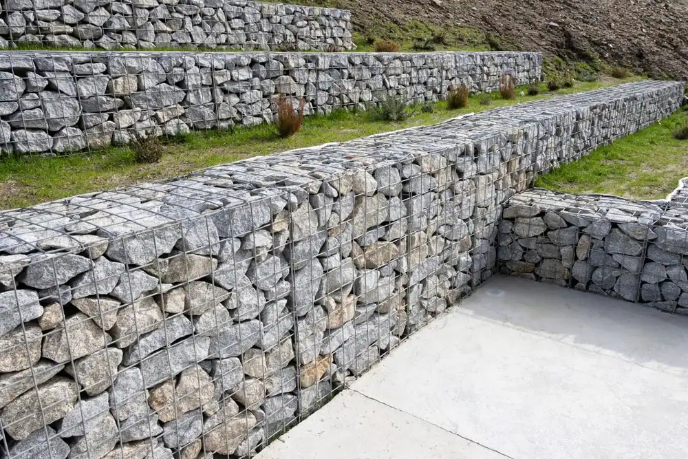 Several gabion retaining walls made of wire cages filled with rocks are arranged in tiers along a grassy, sloped landscape in Hernando County, FL, with concrete pavement in the foreground.