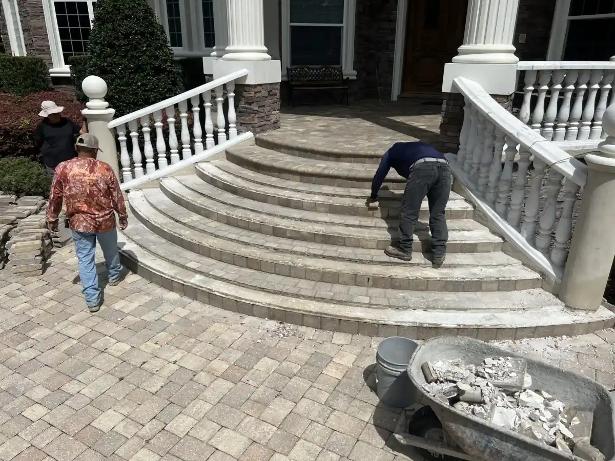 Three workers from a Hernando County landscaper are repairing or installing stone steps in front of a large house with white columns and a balustrade. Debris and tools are visible, along with a wheelbarrow filled with stones in the foreground.