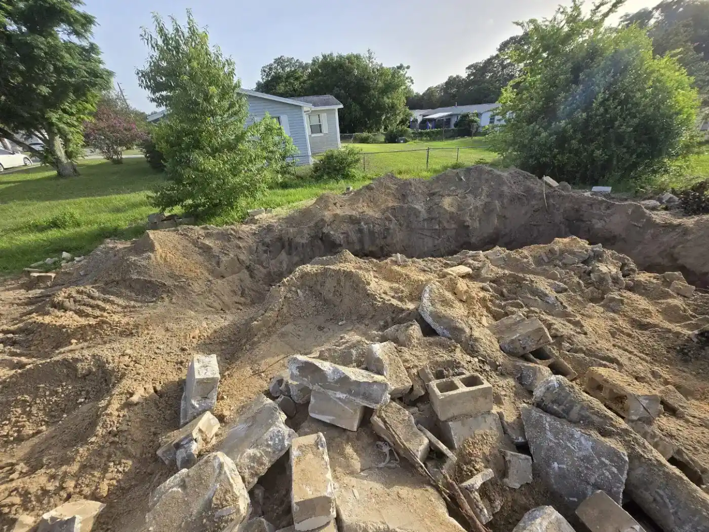 A large dirt hole with piles of rubble and cinder blocks in the foreground, surrounded by green grass, trees, and a light blue house with white trim in the background—typical of rural scenes in Hernando County, FL.