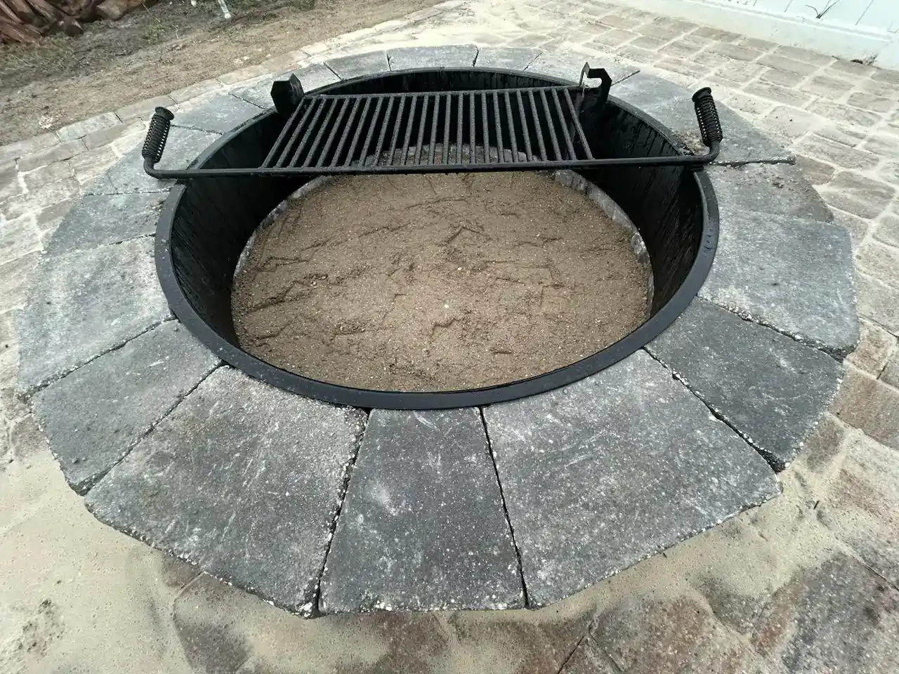 A circular outdoor fire pit in Hernando County, FL, is lined with gray stones and topped with a metal grill grate. Filled with sand instead of firewood or ashes, it sits on a ground paved neatly with stone tiles.