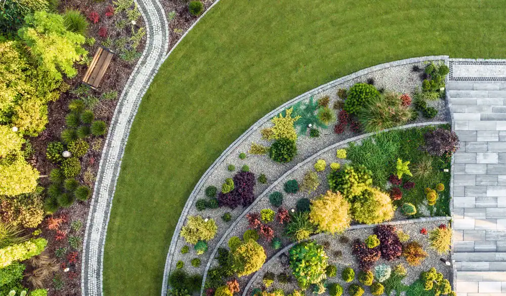 Aerial view of a landscaped garden by FL Landscaper Citrus in Sumter, featuring curved paths, a stone bench, green lawn, and neatly arranged plants in circular, gravel-lined beds next to a paved area.