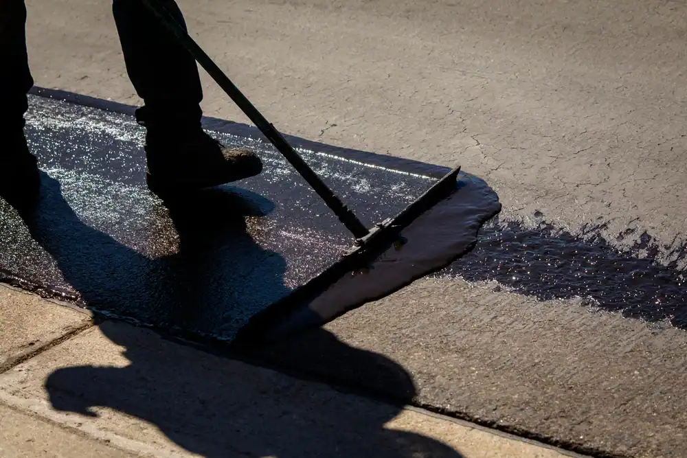 A landscaper in Citrus or Sumter, FL uses a tool to spread black sealant on a cracked asphalt surface, likely as part of a road or driveway repair, casting a shadow on the pavement in bright sunlight.