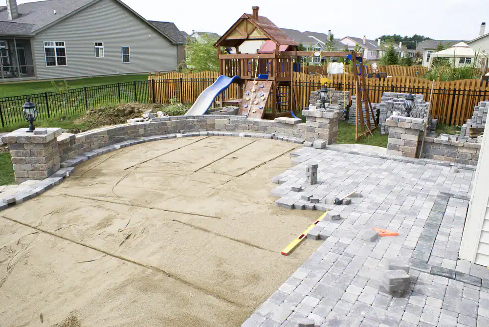 Partially completed backyard patio with paver stones and sand base, tools scattered around, and a children’s wooden playset with a slide and swings in the background, nestled among Hernando County, FL homes with a wooden fence.
