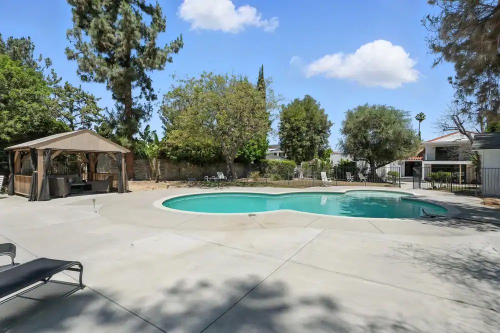 A kidney-shaped outdoor swimming pool surrounded by a large concrete deck, lounge chairs, and a gazebo sits beneath tall trees and blue skies&mdash;perfect for relaxing in scenic Hernando County.