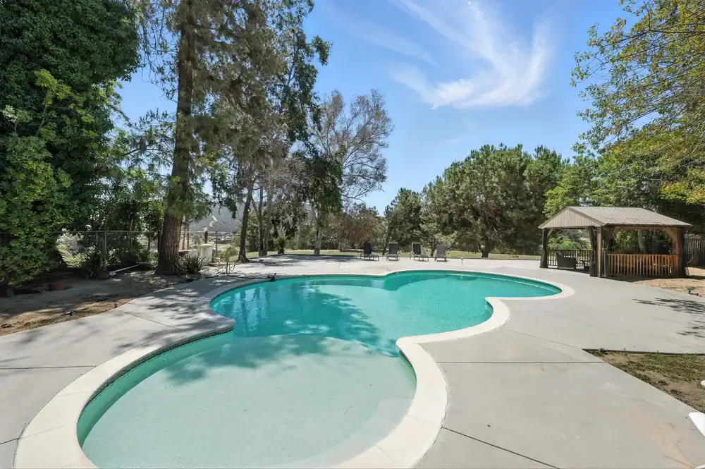 Outdoor swimming pool with a curved shape, surrounded by a concrete deck, tall trees, and a wooden gazebo&mdash;professionally designed by Landscaper Citrus&mdash;in the background under a blue Hernando County sky with wispy clouds.