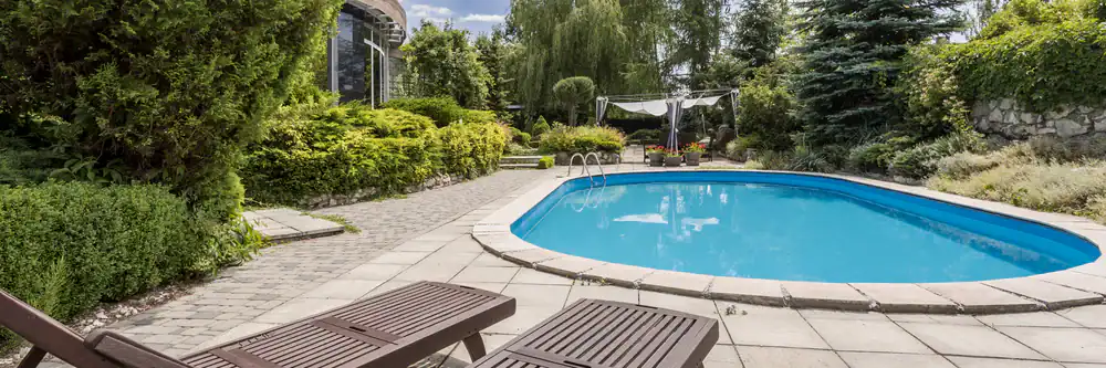 A serene backyard in FL with a curved swimming pool, surrounded by lush greenery, stone pavement, and two brown lounge chairs. Trees and shrubs offer shade, while a canopy sits at the far end&mdash;a perfect Sumter or Hernando County retreat.