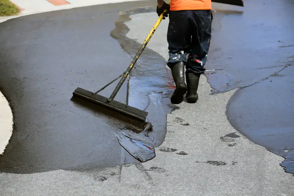 A worker in boots, possibly a landscaper from Citrus or Sumter, spreads black sealcoat over a paved surface with a large squeegee, leaving footprints behind on the wet sealant.