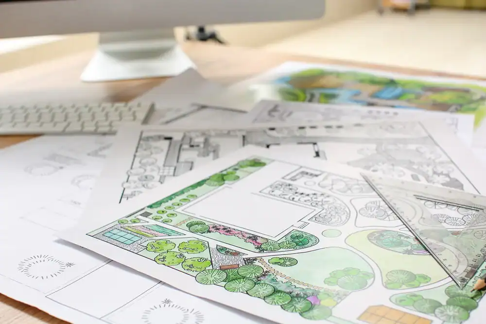 A stack of landscape design plans and sketches showing gardens and green areas is spread out on a desk near a computer keyboard and monitor, illustrating the creative process of a Hernando County or Landscaper Citrus professional.