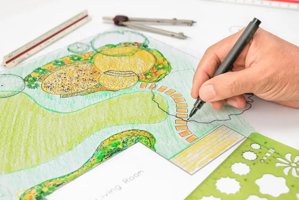 A hand draws and colors a landscape design plan on paper, showing pathways, green areas, and planting beds. Drawing tools, templates, and a scale ruler rest nearby&mdash;just the setup for a skilled Hernando County landscaper.