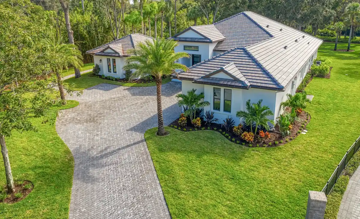 Aerial view of a modern single-story house with a tiled roof, lush green lawn, palm tree, and landscaped garden beds in Hernando County, FL. A paved driveway leads to a detached garage, all surrounded by trees.
