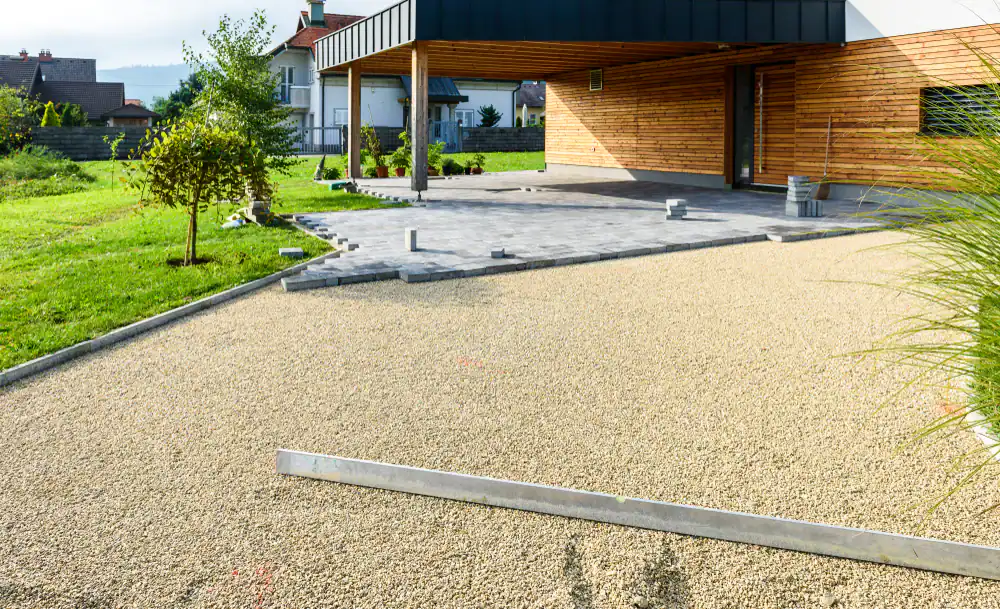 A modern house with wood siding and a covered carport is shown in Hernando County, FL. The driveway is under construction, with gravel and paving stones being installed by Landscaper Citrus. Green grass and small trees border the yard.