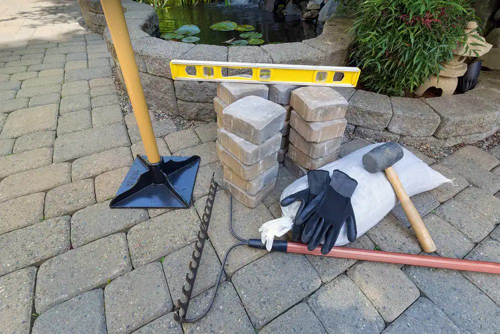 A set of landscaping tools and materials, perfect for any Hernando County, FL project, including a rake, mallet, gloves, paver bricks, a leveling tool, and a bag of sand&mdash;arranged on a stone patio near a pond with water lilies.