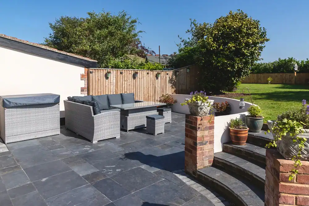 A modern patio in Sumter, FL, with gray stone tiles features outdoor wicker furniture, potted plants, and brick steps leading to a grassy garden&mdash;expertly designed by a local landscaper&mdash;beneath a clear blue sky.