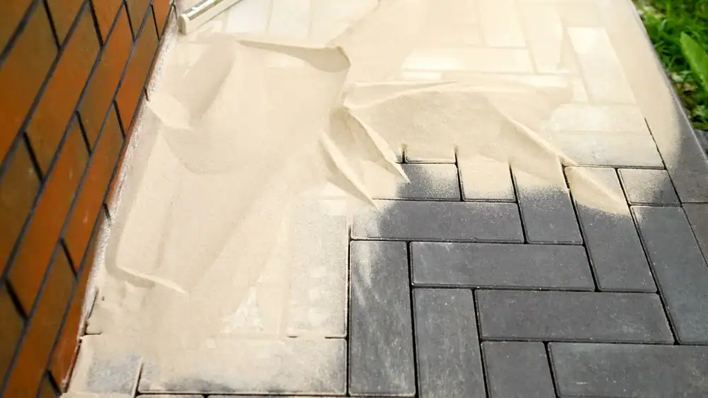 Sanded pavers next to a brick wall, with light-colored sand spread over dark gray interlocking paving stones&mdash;some swept into the gaps and some on the surface&mdash;showcasing detailed work by a skilled Landscaper Citrus, FL.
