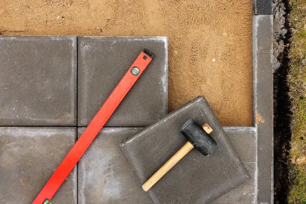 Several dark concrete pavers are being installed on sand in Hernando County. A red spirit level and a rubber mallet with a wooden handle rest on the pavers, with grass and soil visible at the edge.
