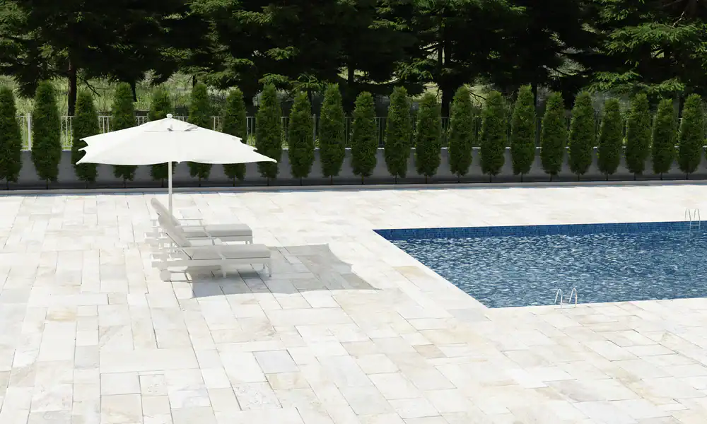 A swimming pool with clear blue water is surrounded by white stone tiles. Two lounge chairs and a white umbrella sit beside the pool, perfectly complemented by tall green shrubs&mdash;an inviting retreat crafted by Landscaper Citrus in Hernando County.