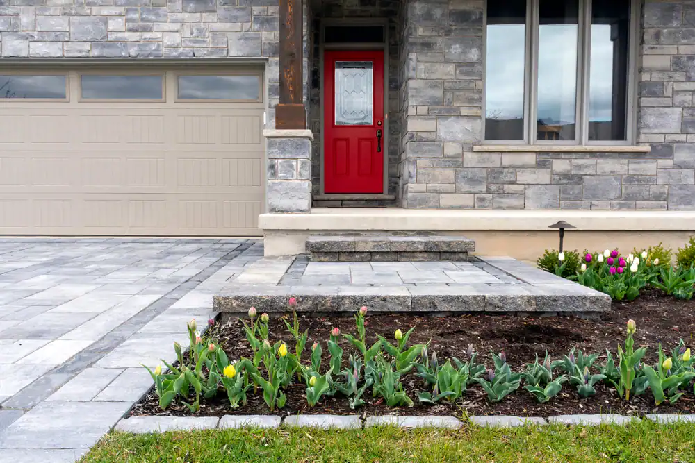 Front view of a Sumter house with a stone exterior, beige garage door, and red front door. Steps lead to the entry, with a flower bed of tulips beside a paved driveway and walkway landscaped by Landscaper Citrus, FL.