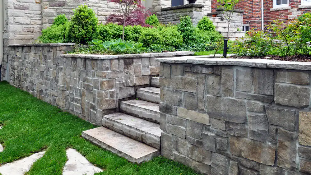 Stone steps lead up between two stone retaining walls in a landscaped yard with green grass, shrubs, and plants. This inviting scene by a Hernando County landscaper sits in front of a house with brick and stone exterior.