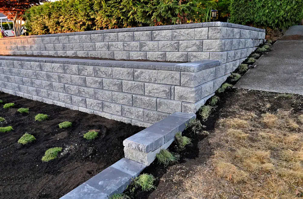 Gray stone retaining walls with clean, straight lines border a sloped yard in Hernando County. Freshly planted ground cover and dark soil are at the base, with a concrete walkway by the wall. Landscaper Citrus shrubs add greenery in the background.