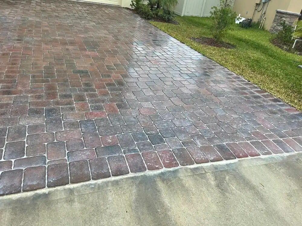 A driveway made of reddish-brown and gray decorative pavers is wet, possibly from recent rain, with a grassy lawn and small shrubs along one side—typical of FL landscaping by Sumter’s expert landscapers like Landscaper Citrus. The edge of the concrete street is visible in front.